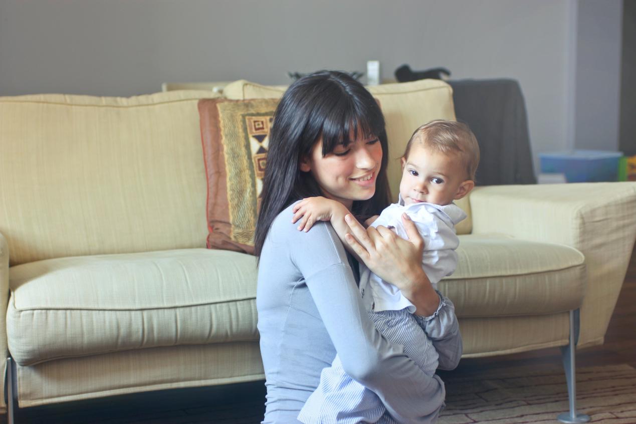 A woman smiles at the baby she is holding, while sitting in front of a couch.