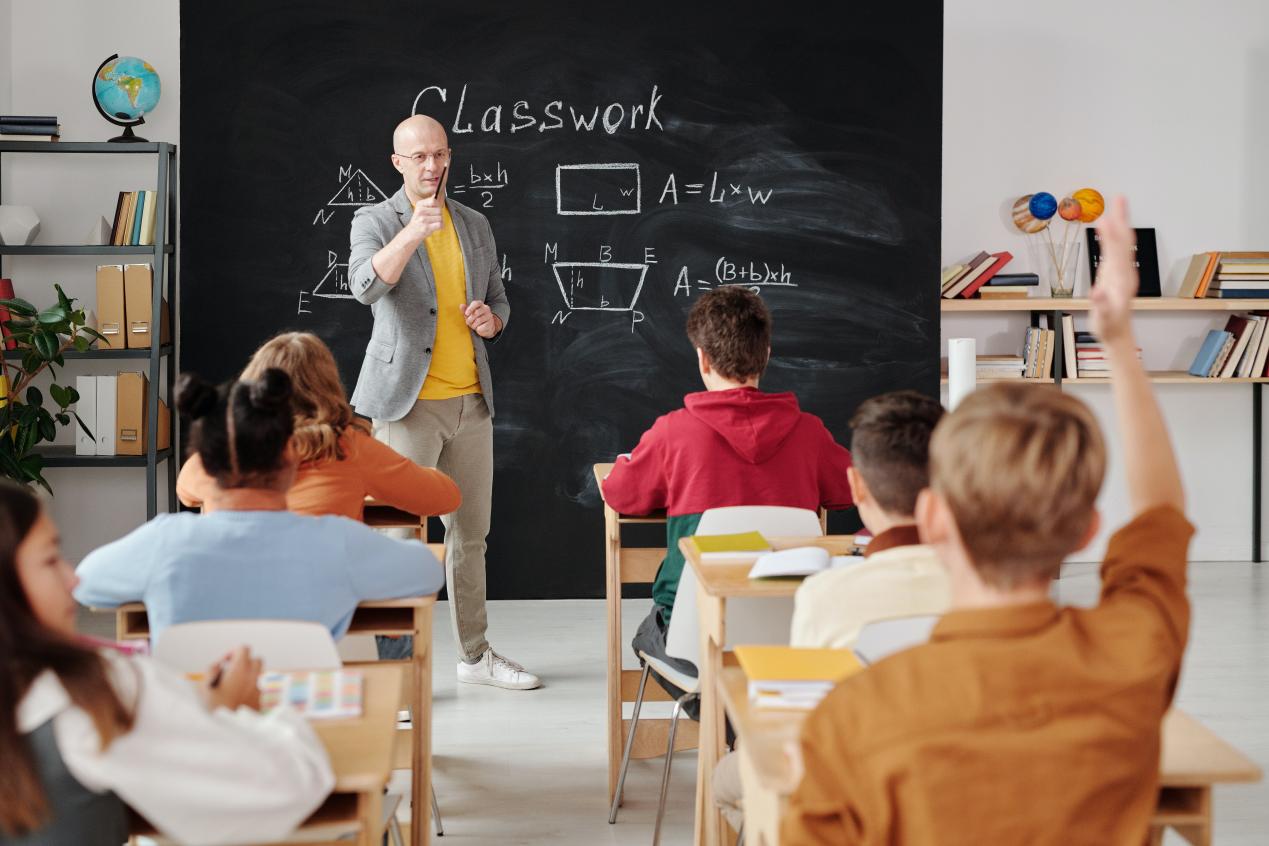 A teacher stands in front of a chalkboard, raising his pencil to call on a student raising his hand.