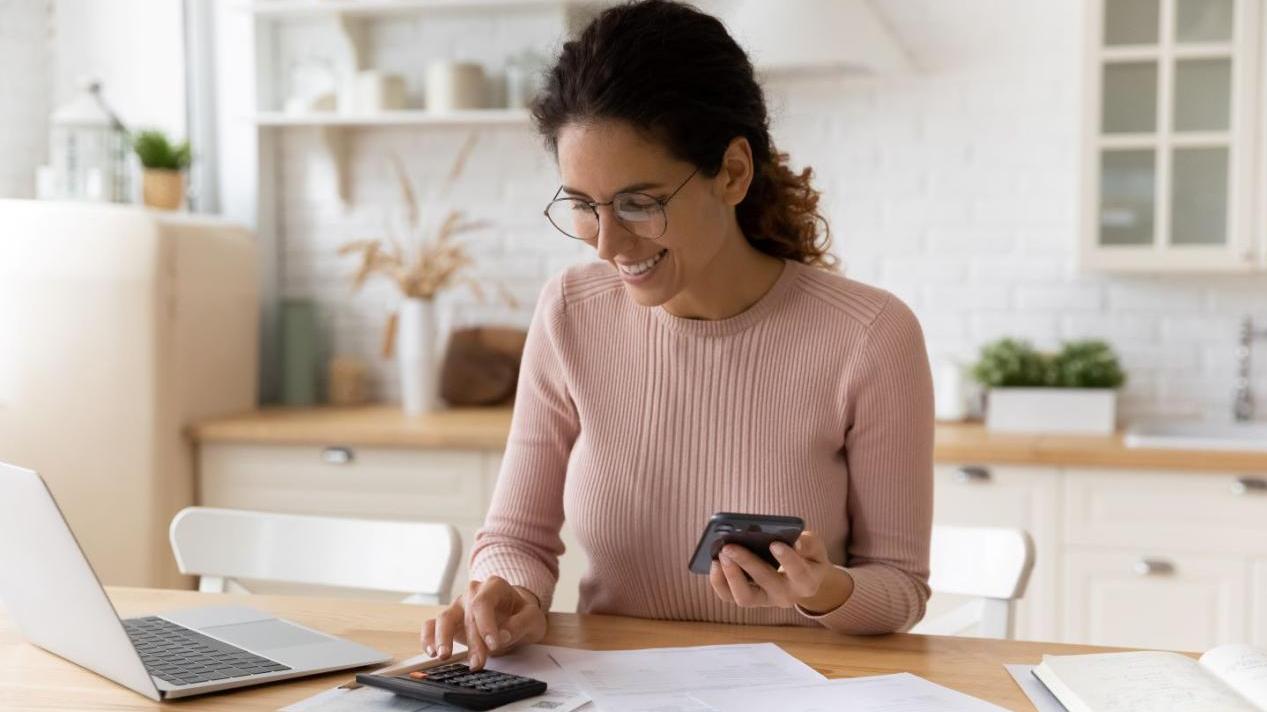A woman uses a calculator while looking at tax documents and a phone.